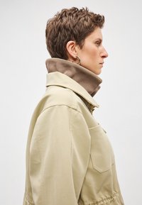 Μπεζ πουκάμισο με ψηλό γιακά, διαθέτει καφέ εσωτερική επένδυση, πλευρική τσέπη και υφή υφάσματος. Κλασικός σχεδιασμός με εφαρμοστή γραμμή.