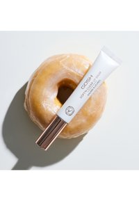 En klar læbebalsam tube med en hvid krop og en skinnende roseguld kap, placeret ovenpå en glasur donut på en hvid baggrund.