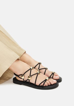 Černé sandály s pásky a plochou podrážkou, s pozlacenými kruhovými ozdobami na páskách. Design zahrnuje minimalistický styl s otevřenou špičkou.