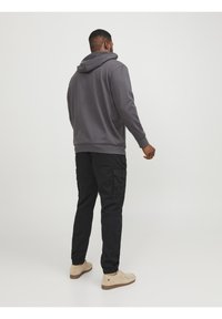 Grijze hoodie met een ruime capuchon, gecombineerd met zwarte cargobroeken met zijzakken. Het model draagt lichtbruine schoenen en staat met de rug naar de camera.