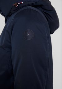 Mörkblå hooded jacka av slät tyg, med en rund logotyp på ärmen och kontrasterande dragsnoddar vid huvan.