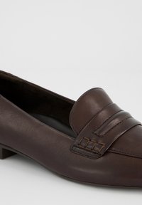 Mocassin femme en cuir marron foncé avec un petit talon et une bride décorative avec détail cousu sur un fond uni.