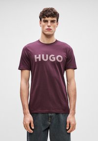 Burgundia T-särk, millel on julge, heledat värvi "HUGO" tekst. Lühikesed varrukad, ümar kaelus ja pehme kangas. Kanda tumedate denimpükstega.