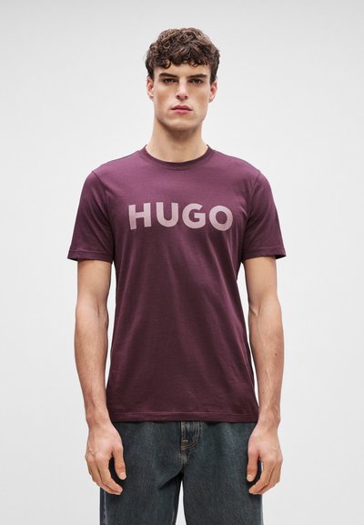 Бордо тениска с ярък, светъл надпис "HUGO". Къси ръкави, кръгло деколте и мека материя. Съчетава се с тъмносини дънки.
