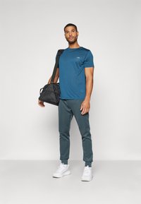 Blå strukturert trenings-T-skjorte, grå joggebukser med hvite sidestriper, og hvite sneakers. Svart treningsbag hengende over skulderen.