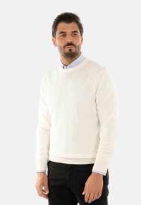 Maglione bianco a maglia con scollo rotondo, maniche lunghe e polsini a coste, abbinato a una camicia azzurro chiaro con colletto sotto.