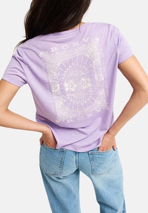 Femme aux longs cheveux foncés porte un t-shirt lavande avec un motif floral et solaire blanc complexe et un jean bleu clair, mains dans les poches arrière.