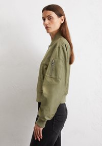 Olivgrüne Bomberjacke mit einer Reißverschlusstasche am linken Arm, gerippten Bündchen und Saum sowie einer glatten, leicht glänzenden Textur.