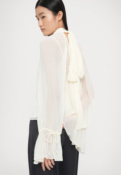 Noor naine, kes istub profiilis ja kannab läbipaistvast valgest õhukesest pluusist koos pikkade lehvivate varrukatega ning suure sõlmega vöökohal tagant.