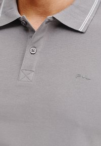 Szary polo z teksturowanej tkaniny, z dwoma guzikami, ściągaczem i delikatnymi białymi paskami. Wyhaftowane logo.