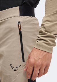 Pantalon beige avec poche zippée noire et petit logo visage d’animal près de la taille ; main d’une personne et manche longue beige visibles.