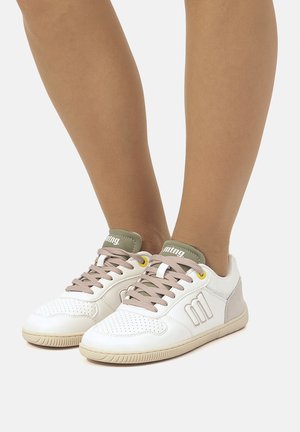 Benen met witte lage sneakers met lichtroze veters, olijfgroene tong en beige zool op een effen lichte achtergrond.