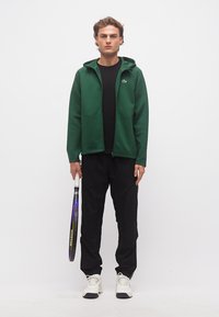 Νέος άνδρας με πράσινο φούτερ με φερμουάρ, μαύρο παντελόνι και λευκά αθλητικά παπούτσια, κρατώντας ρακέτα τένις δίπλα του σε απλό φόντο.