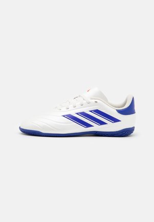 Zapatilla deportiva blanca con acentos azules, con franjas laterales, parte superior sintética lisa, suela texturizada y cierre con cordones.