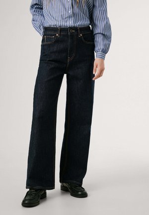 Jeans en denim bleu foncé à jambes larges avec des coutures orange, poches avant et fermeture à bouton. Associés à une chemise rayée bleue et des chaussures noires.