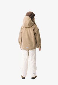 Tan fleecejakke med hætte, parret med cremefarvede bukser og brune fodtøj. Stoffet har et tekstureret, hyggeligt udseende.