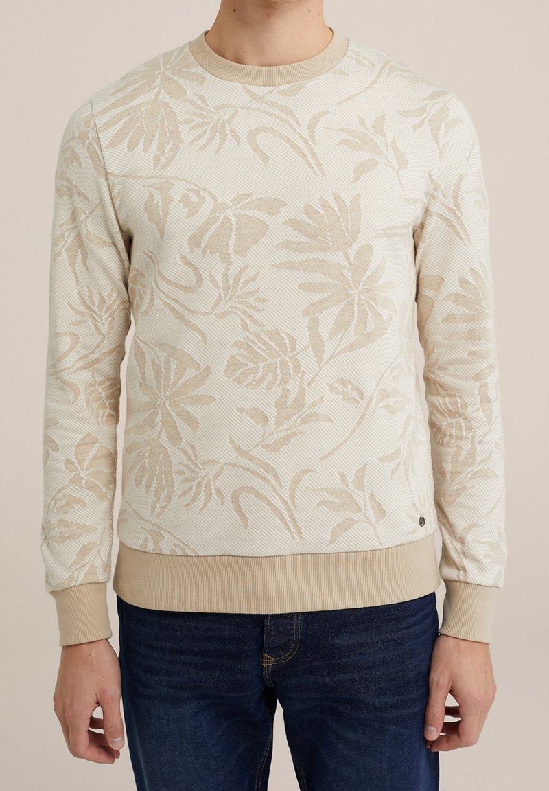 Beige och vit blommönstrad långärmad tröja på en person som bär mörkblå jeans, stående mot en neutral bakgrund.
