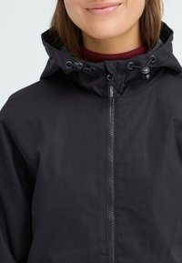Svart vanntett jakke med hette, eksponert glidelås og snor. Glatt tekstur med minimalistisk design. Merkeetikett synlig.
