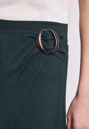 Donkergroene rok met een grote ovale zwarte gesp in de taille, gedragen met een witte top, waarbij een deel van een arm zichtbaar is.