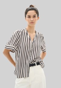 Gestreiftes Hemd in hellbeige und marine blau, kurzärmlig, V-Ausschnitt, strukturiertes Material. Kombiniert mit hoch sitzenden weißen Hosen und einem schwarzen Gürtel.