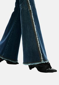 Tumesinised hõlpsasti alt laienevad teksad, millel on narmendavad servad ja pruunid õmblused külgedel. Sobitatud teravate mustade kontsakingadega.