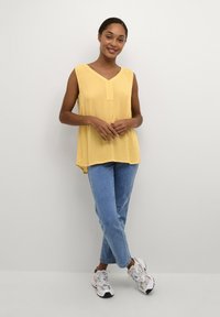 Yellow sleeveless top with a V-neck and a button detail, paired with light blue jeans and white sneakers. Smooth fabric with a relaxed fit.