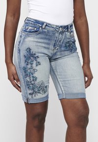 Shorts en denim bleu clair avec broderies florales en bleu foncé et vert. Comprend des ourlets roulés et un design traditionnel à cinq poches.