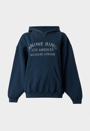 Jeune personne portant un sweat à capuche sombre oversize avec des inscriptions, un jean bleu ample et des chaussures pointues noires, debout devant un fond uni.