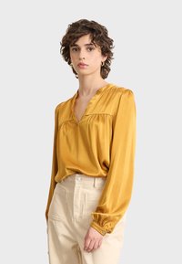 Mujer con cabello corto y rizado que lleva una blusa de manga larga mostaza y pantalones de tiro alto beige, de pie contra un fondo sencillo.
