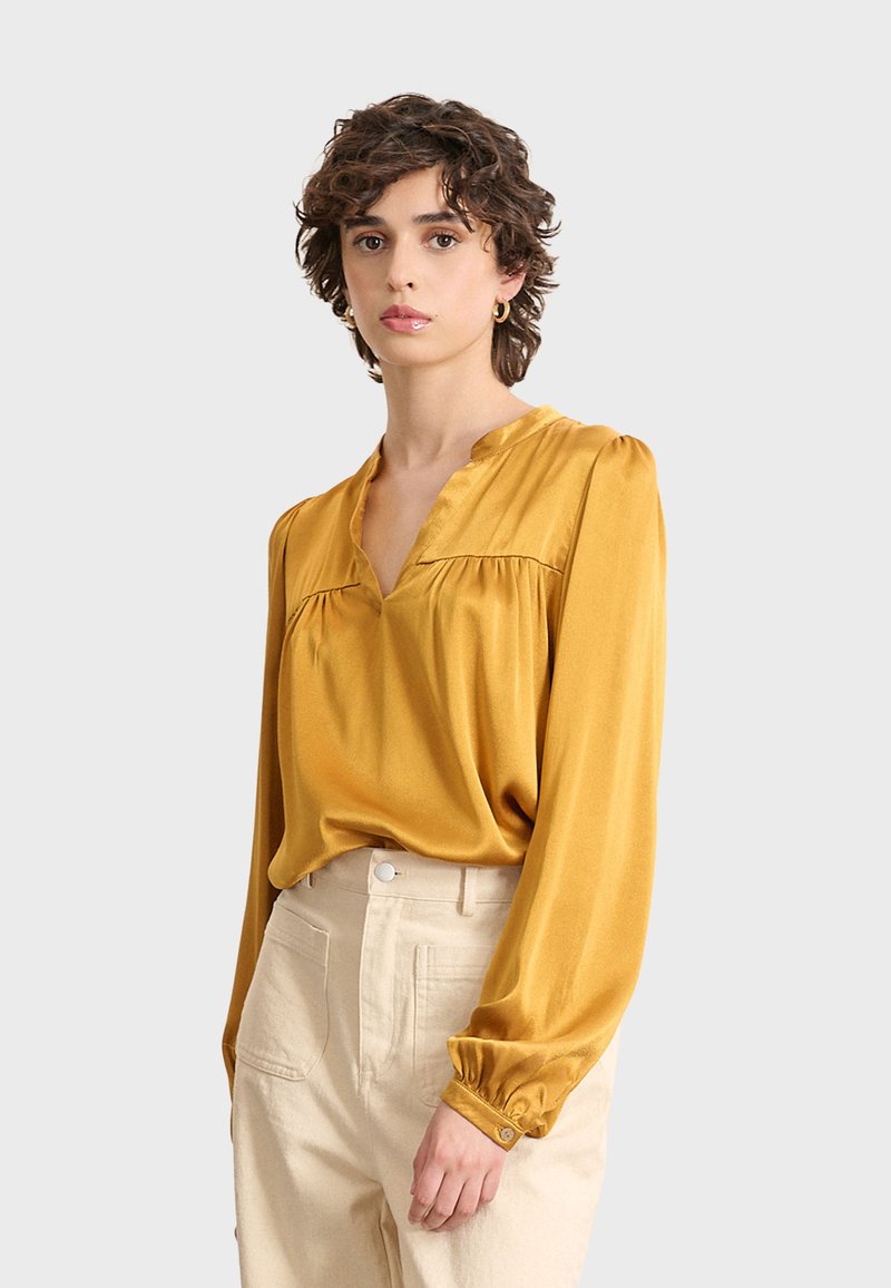 Mujer con cabello corto y rizado que lleva una blusa de manga larga mostaza y pantalones de tiro alto beige, de pie contra un fondo sencillo.