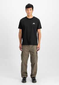T-shirt en coton noir avec un petit logo blanc, associé à un pantalon cargo olive avec des poches latérales, et des chaussures noires avec une finition lisse.