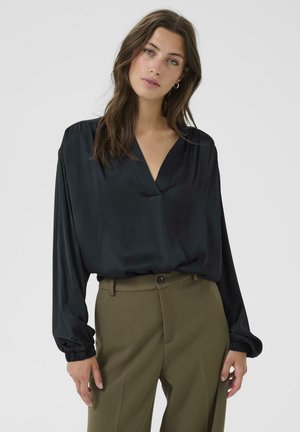 Camicia di satin nera con scollatura a V e maniche lunghe; presenta polsini arricciati e una vestibilità morbida, indossata con pantaloni su misura di colore verde oliva.