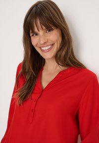 Blusa rossa con una texture morbida, scollatura a V e dettagli con bottoni. Le maniche lunghe e il tessuto liscio arricchiscono il design.