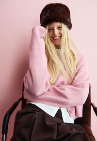 Cardigan en laine rose clair avec deux boutons, superposé à une chemise blanche ; jupe plissée marron ; chapeau en fourrure foncé ; assise sur une chaise marron.
