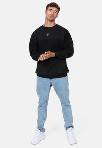 Schwarzer Sweatshirt mit strukturiertem Design und rechteckigem Logo-Patch, kombiniert mit hellblauen Jeans und weißen Sneakers.