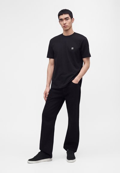 Jeune homme debout, une main dans la poche, portant un t-shirt noir avec un petit logo blanc, un pantalon noir et des baskets noires avec des semelles blanches.