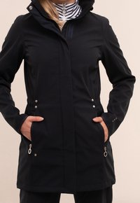 Zwarte, zachte shelljack met een capuchon, twee zijzakken met rits en accenten van zilver hardware. Het heeft een gladde textuur en een gestroomlijnd ontwerp.