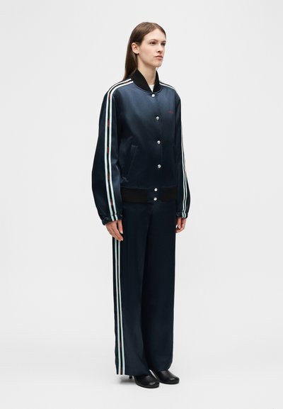 Γυναίκα όρθια φορώντας σατέν σακάκι και παντελόνι σε ναυτικό μπλε με ανοιχτού γαλάζιου ρίγες στα μανίκια και τα πόδια του παντελονιού, μαύρα παπούτσια.