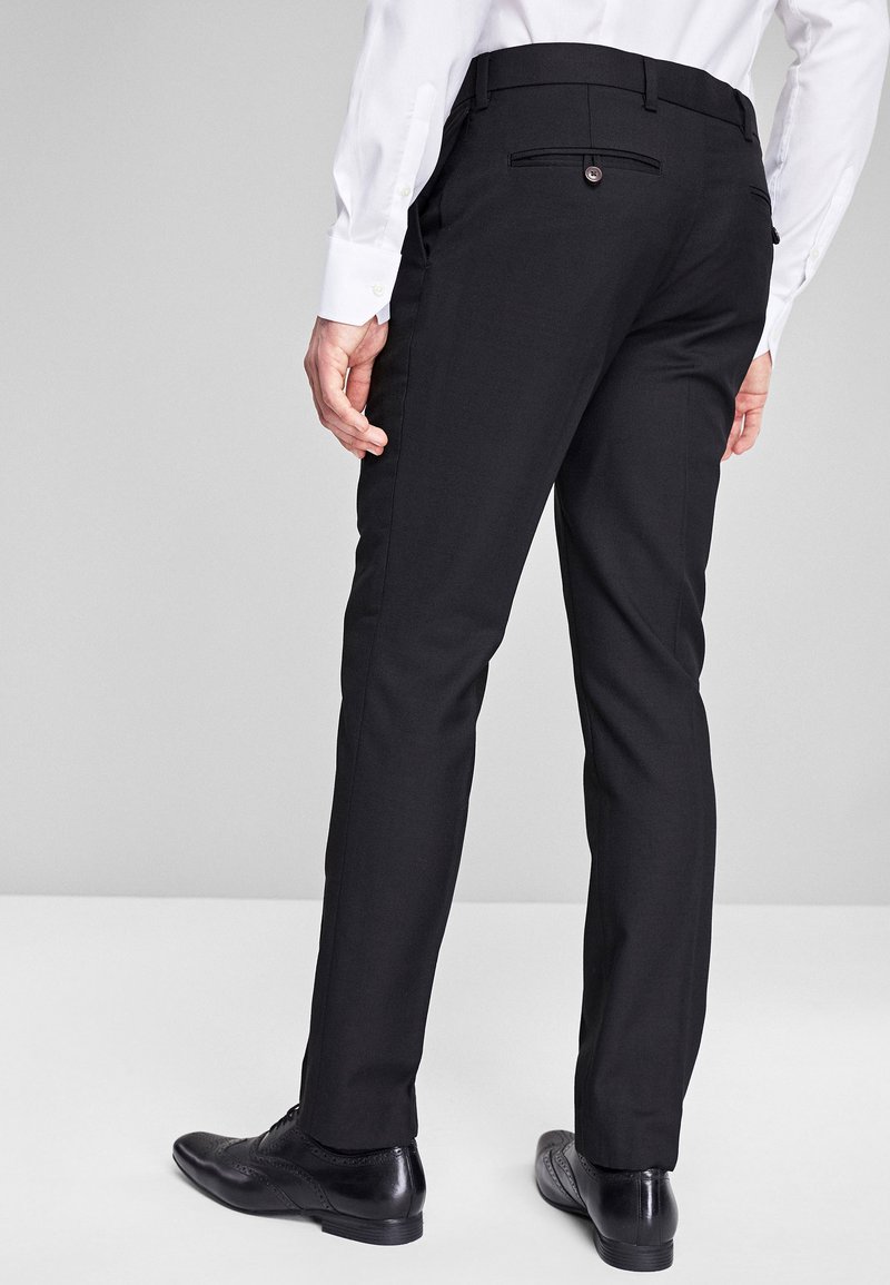 Uomo che indossa pantaloni neri su misura, una camicia bianca e scarpe eleganti di pelle nera, in piedi contro uno sfondo grigio chiaro uniforme.