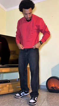 Red sweatshirt, black jeans, and black-and-white sneakers. Features a round neckline and textured fabric. Background includes a TV and standalone fireplace.