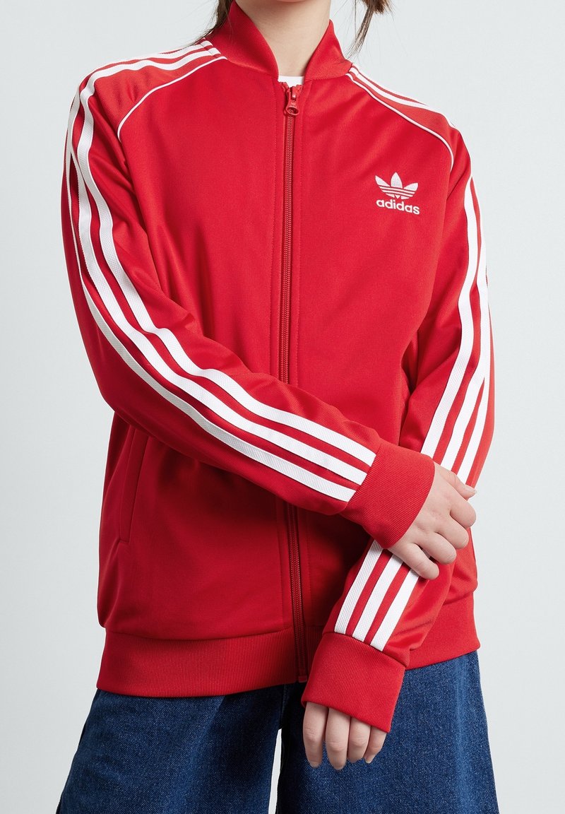 Červená sportovní bunda na zip s bílými třemi pruhy po stranách rukávů. Žebrované manžety a límec, vyrobeno z hladké tkaniny. Logo Adidas na hrudi.
