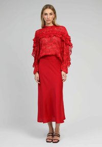 Haut en dentelle rouge avec des manches volantes associé à une jupe midi rouge ajustée, présentant une texture lisse et un subtil éclat. Talons noirs à lanières.