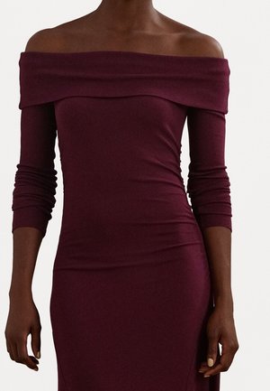 Donkere huidtint, gekleed in een nauwsluitende, off-shoulder, lange mouwen bordeauxrode jurk met een geplooide halslijn en zichtbare natuurlijke nagels.