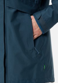 Giacca impermeabile blu navy con una texture liscia, caratterizzata da una mano in una tasca laterale, polsini regolabili con velcro e un piccolo dettaglio con etichetta verde.