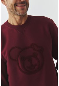 Sudadera burdeos con un motivo de oso texturizado en el frente. El material es suave, con un cuello redondo y puños de canalé.