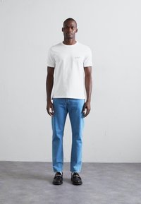 Valge puuvillane T-särk helesinise broderiga logoga, ühendatud helesiniste teksade ja mustade elegantsed jalanõudega, seistes neutraalse tausta ees.