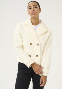 Manteau crème à double boutonnage en tissu texturé, avec de grandes boutons dorés et un large col. Les manches sont légèrement retroussées.