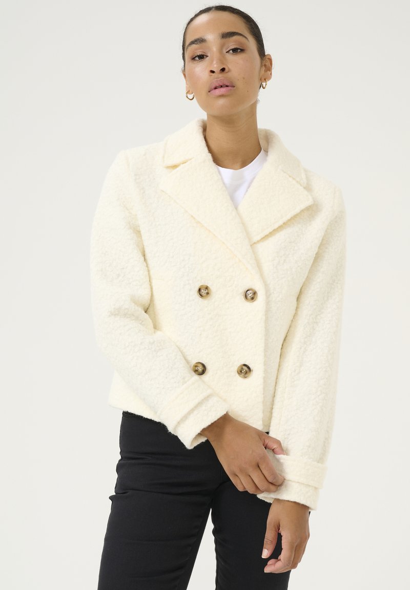 Manteau crème à double boutonnage en tissu texturé, avec de grandes boutons dorés et un large col. Les manches sont légèrement retroussées.