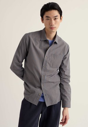 Mężczyzna ubrany w szaro-brązową kratowaną koszulę na guziki, założoną na niebieski t-shirt, stoi z jedną ręką za plecami na jednolitym tle.