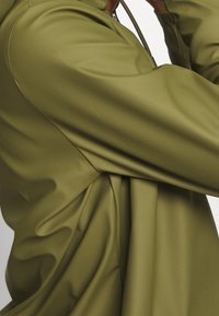 Vêtement en tissu lisse vert olive, à la coupe oversize, présentant une texture froncée et un cordon à nouer au col.